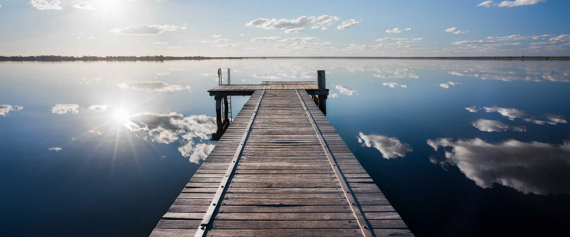A wooden dock extends over a clear, blue lake on a sunny day. Puffy white clouds are reflected on the surface of the water.