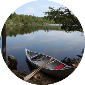 A calm lake with a metal canoe pulled up to a rocky shore. A red lifejacket and wooden oars sit in the canoe. 