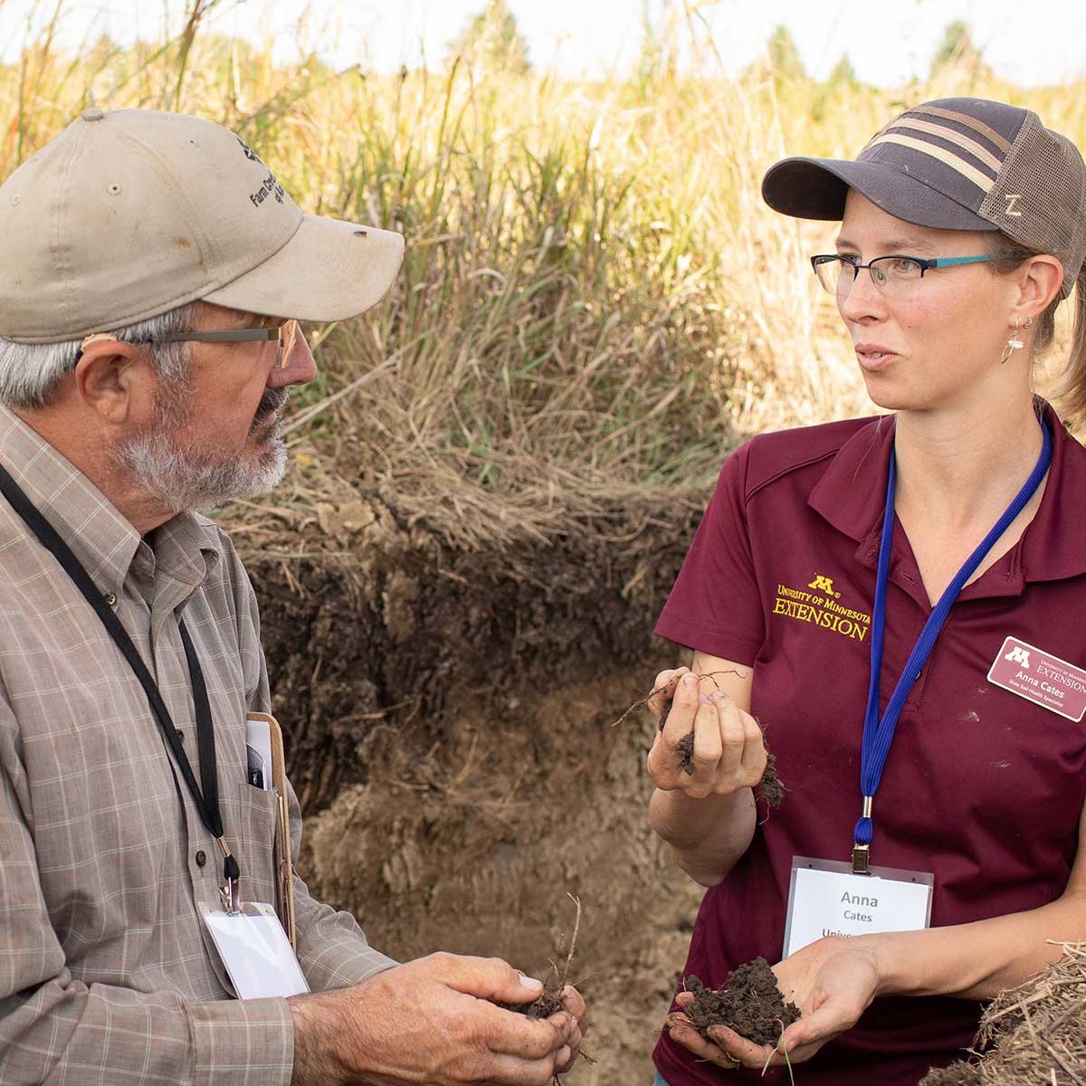 An older white man wearing a tan baseball cap and glasses holds a handful of soil while listening to a younger white woman with a handful of soil.. She is also wearing a ball cap and glasses, also an U of M Extension name badge.