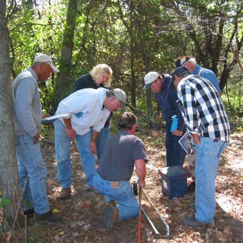 Seven individuals stand outside in a forested area looking at exposed pipes in the ground.