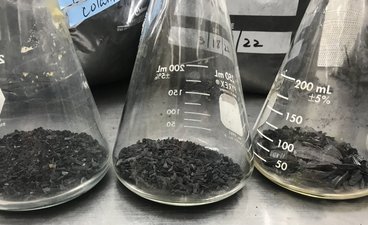 Three Erlenmeyer flasks on a metal lab bench contain biochar