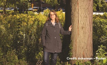 Woman in a black raincoat stands next to a tree on the St. Paul campus